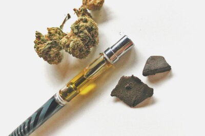 Comparison of cannabis forms showing dried flower buds, a liquid vape pen cartridge, and solid chunks of traditional hashish on a white background.